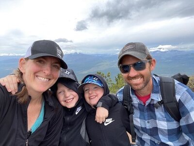 family photo in an open location with mountains. the social media strategist is wearing a black sports cap and waterproof jacket. in the center are two children wearing caps and jackets with hoods. and the man on the left side is wearing dark glasses with a blue plaid shirt