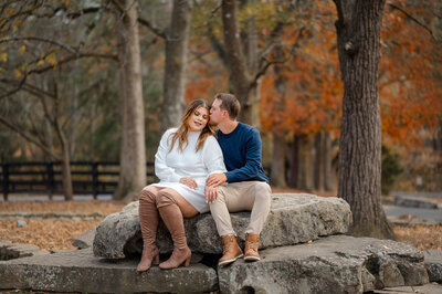 Engaged couple sit together on a rock at Sims Lake Park while her fiance kisses her on the side of the head and she closes her eyes to enjoy the moment together