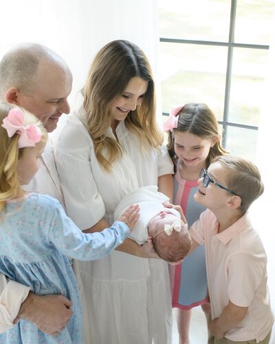 Parents and siblings gather around, smiling at their swaddled newborn in soft natural light. A loving family session by Moments in Light Photography.