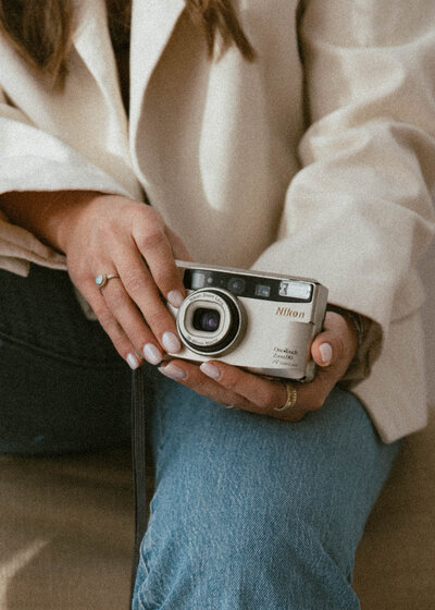 A woman holding camera sitting on a chair wearing a cream blazer and jeans