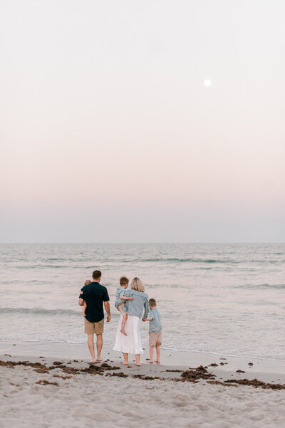 Family of five standing down by the water in Cape Canaveral, Florida at sunset looking out at the ocean.
