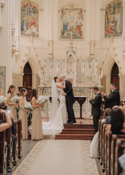 Bride and groom share their first kiss at the altar of a grand, ornate church while their bridal party looks on.