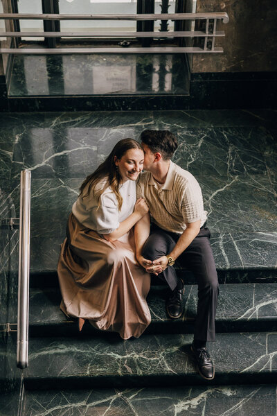 Couple sitting on a stairwell, with the man leaning in to whisper in his fiancée's ear.