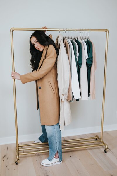 Woman with long, brown hair wears a camel-colored trenchcoat that hangs from a hanger on a clothing rack.
