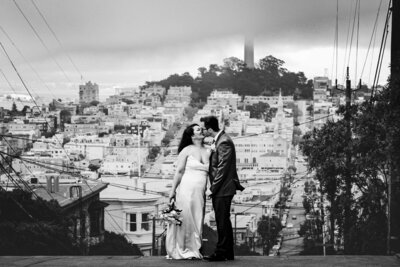 a black and white image of a bride and groom kissing on a street in san francisco with coit tower and hills of buldings in the background. the sky is foggy. the bride holds a bouquet and wears a white satin gown. The groom is in a dark suit.