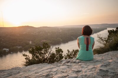 woman in a green shirt sitting down on a rocky ledge watching the sunset