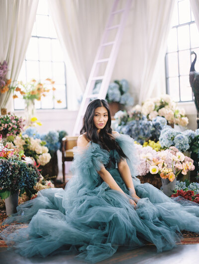 Model in blue tule dress sits on the floor surrounded by flowers