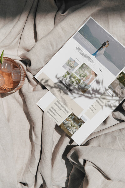 A magazine or brochure with wedding-themed photos lies open on beige fabric next to a glass of iced drink. Branch shadows fall across the scene, evoking elegant inspiration—perfect for exploring wedding photography SEO or seo for creatives.