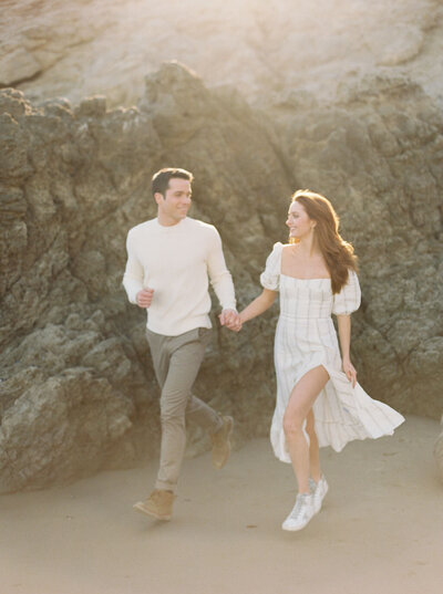 engaged couple running on beach in front of cliff
