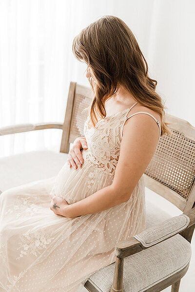 A pregnant woman in a lace dress sits on a wooden chair, gently holding her belly and looking down. With sheer curtains in the background, this serene moment is beautifully captured by Destin maternity photographers.