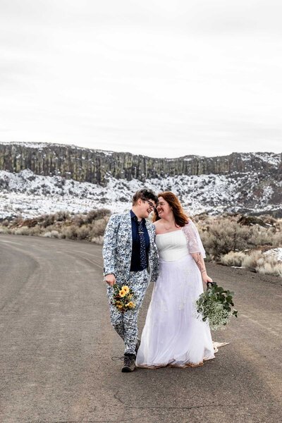 Two brides are walking side by side with their heads close together both holding bouquets. One is wearing a floral blue suit and the other is wearing a white dress. They are walking along a gravel road with a snowy rocky background