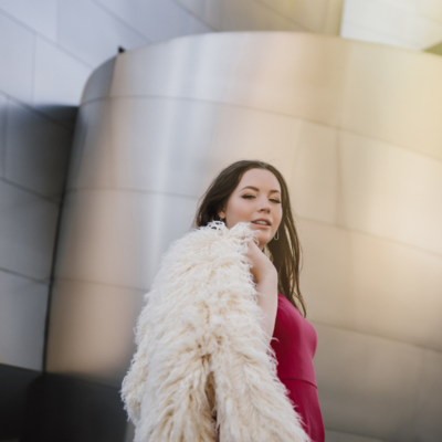 high school senior girl in pink dress holding a furry white coat over her shoulder looking at camera in front of disney music hall