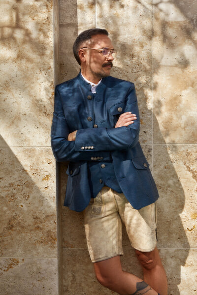 Mann in modernem Trachtenoutfit mit blauem Blazer, traditioneller Hirschlederhosen und blauer Trachtenweste. Das Outfit stammt vom Designerlabel Monaco Spatzl und vereint traditionelle Elemente mit modernem Stil.