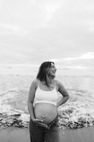 A pregnant woman wearing a white tank top and a pair of jeans during a maternity photoshoot at the beach in christchurch with lucy jane photography