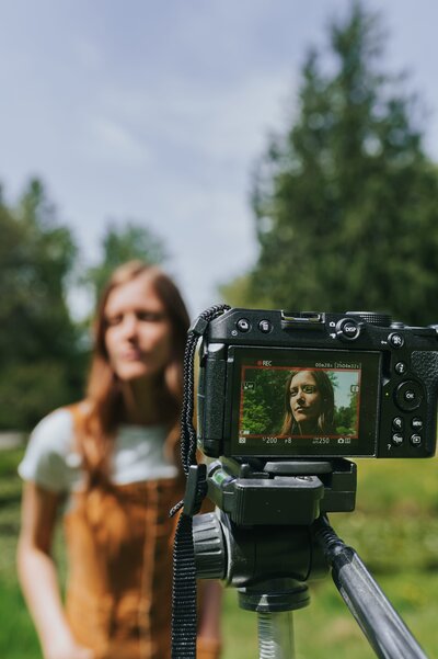 Camera sits on tripod shooting a picture of woman standing outdoors in a mustard-colored jumpsuit and white short-sleeve t-shirt.