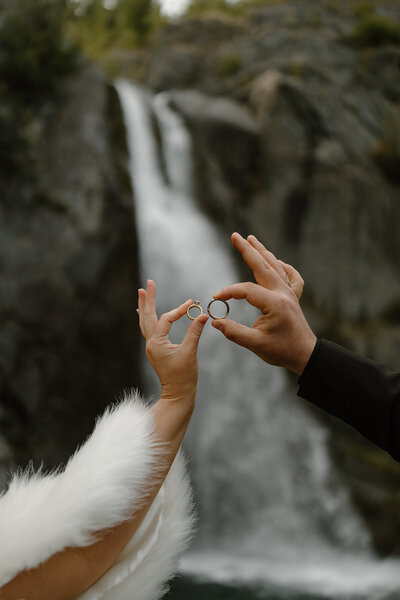 Two hands hold wedding rings up with a waterfall in the background.