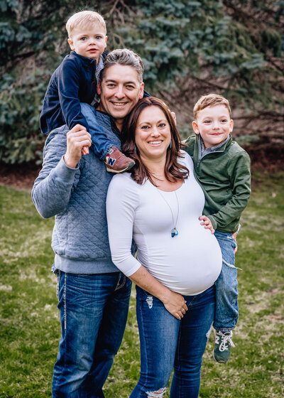 A family portrait featuring a pregnant woman, her partner, and two young boys. The man holds one boy on his shoulders while the other boy stands close to his mother. They are all smiling and dressed casually, standing in a lush green outdoor setting.