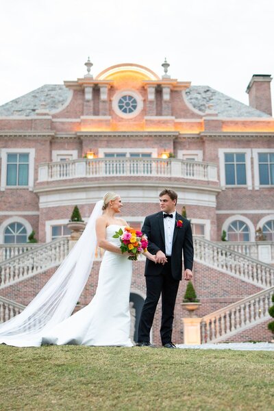Bride and groom posing outside grand mansion with large pink and coral bouquet