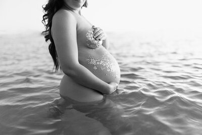 A black and white photo of a pregnant belly at the beach at sunset during a maternity session at St. Andrews State Park in Panama City Beach, FL.
