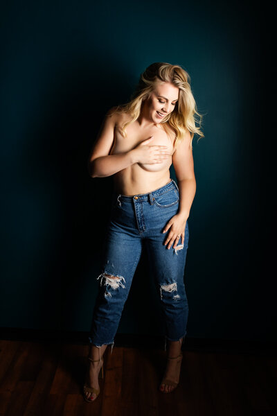 A blonde woman wearing jeans smiles while covering her topless chest with her arm