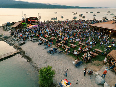 Aerial view of a large crowd gathered for a lakeside concert at sunset, with boats anchored nearby and a stage set up along the shoreline, captured by Joseph Mayden.