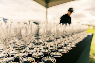 Rows of clean wine glasses on a table under a canopy with a server in the background, captured by Joseph Mayden as part of professional event photography coverage.