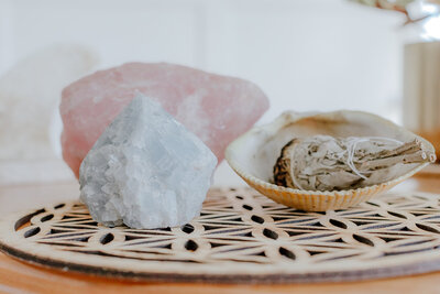Celestite and rose quartz crystals with sage bundle in a seashell arranged on a decorative wooden mat in a light, airy room - Life Coach - Susie Schumacher