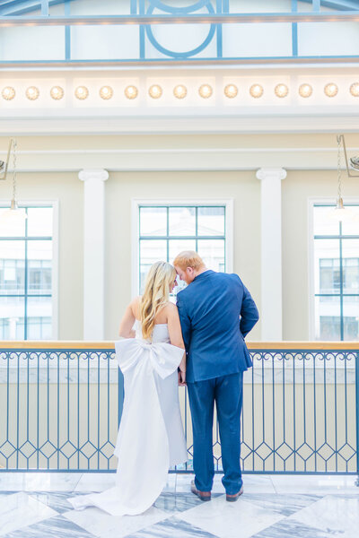 A groom wearing a blue suit and a bride wearing a long white satin dress with a big bow on the back facing away from the camera lean in forehead to forehead