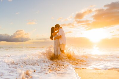 young engaged couple kisses on hawaii beach as shorebreak washes over them in maui