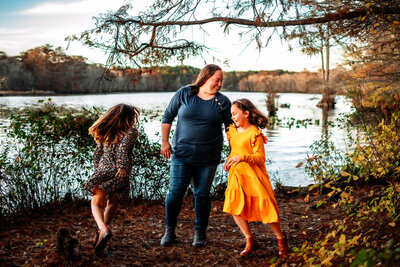 A mom is standing in the middle smiling while looking down while two little girls are running around her with their hair blowing in the wind while wearing flowy dresses. Theres leaves on the ground and water in the background and a tree branch above them.