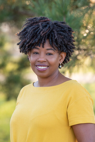 An African American woman in a yellow dress smiling and posing for her headshot session at a park.