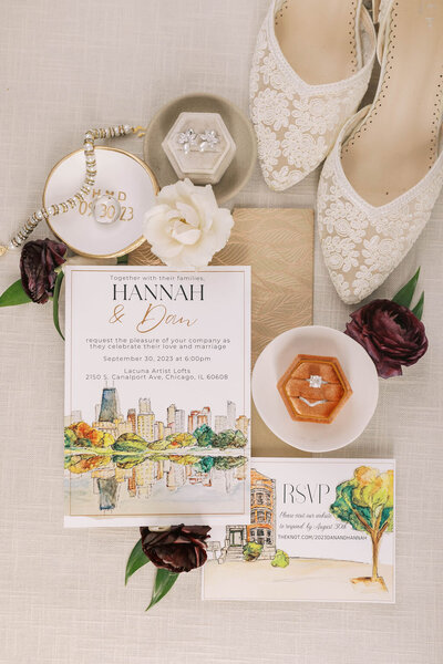 Flatlay displays colorful watercolor wedding stationery with burgundy roses and elegant engagement ring and wedding band.