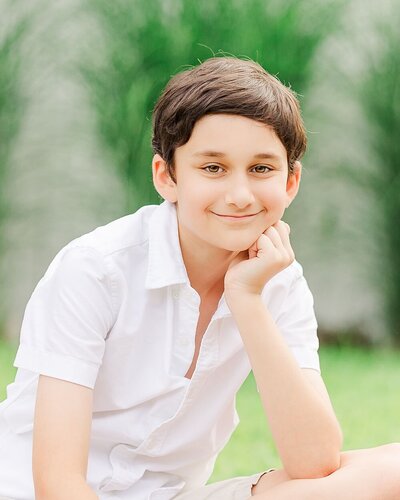 Boy with a white shirt sitting with his hand on his chin outdoors.