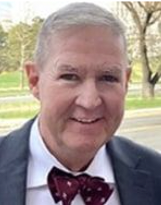 Smiling mature man wearing a dark suit, white shirt, and red patterned bow tie, standing outdoors with trees and a road in the background.