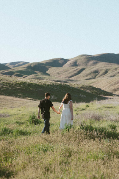 Couple holding hands while walking through a sunlit field in the Idaho foothills during an engagement session