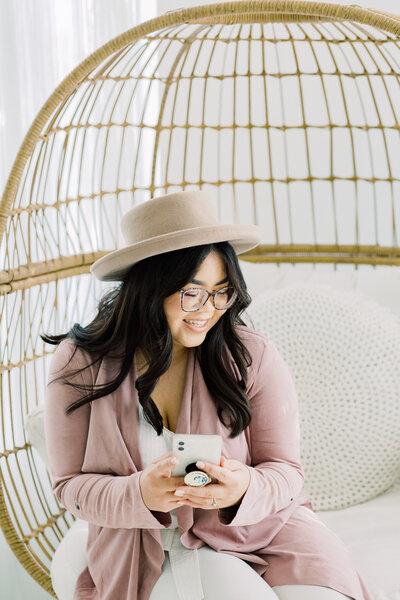 Donna from Donna and Matthew Photography on her phone for a fun playful headshot for her brand images sitting on a rattan egg chair in an all white studio captured in natural light.