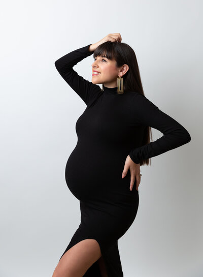 Expectant mom in black form-fitting dress is standing side-profile to the camera. One hand is resting at the small of her back, the other one is atop her head. She is smiling towards the light. Captured by top Brooklyn Maternity Photographer Rochel Konik.