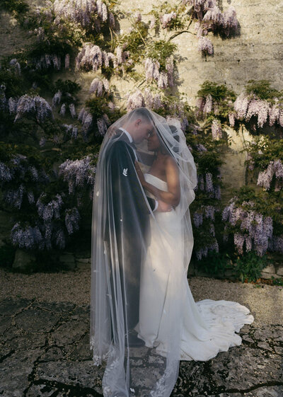 Bride and groom standing in front of sunlit wisteria at Caswell House