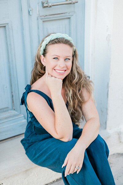 Ciara, assistant planner at Lefkas Weddings, smiling in a relaxed portrait while seated on steps in front of a light blue door