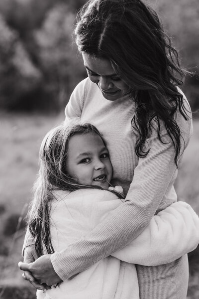 a black and white photo of mom looking down at and hugging her daughter and the daughter looking at the camera smiling