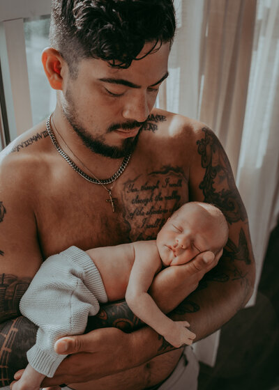 shirtless tattooed dad hold tiny newborn son in his arms during a newborn session near denver colorado
