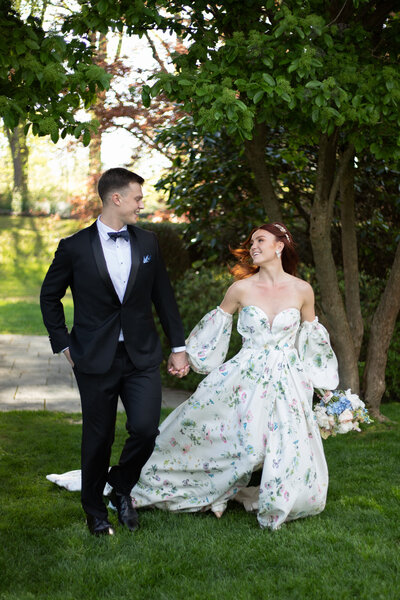 Rhode Island Wedding Photographer | Kelsey Sheehan Photography Rhode Island Wedding Photographer | A bride in a floral gown and groom in a tuxedo joyfully hold hands, walking on grass under trees. It is a sunny day, evoking happiness and romance.