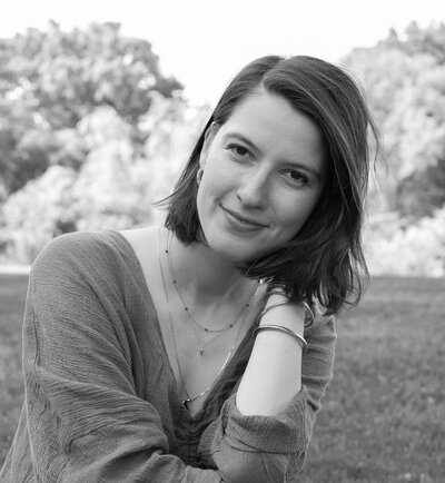 Black and white photo of woman with short brown hair sitting outdoors smiling softly at the camera with trees in the background.