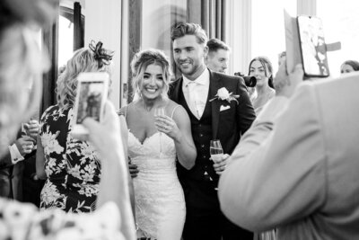 candid black and white photo of a bride and groom holding champagne glasses and surrounded by their cheering guests as they walk into their reception at Hampton Manor