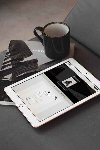 Tablet displaying Emilia Showit website booking page beside a black coffee mug and modern magazine on gray fabric chair
