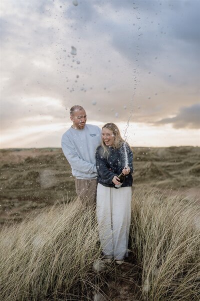 A couple stand on a sand dune at an Aberdeenshire beach. The stand beside each other and are photographed spraying a champagne spray.