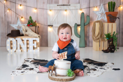 With one hand holding his cake topper and cake in between his legs, overalls dressed baby sits for his portraits with ANE Photography.