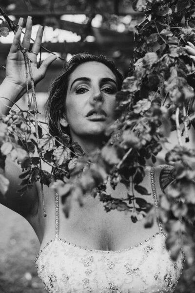 Woman standing behind branches with sultry look on her face in wet wedding dress