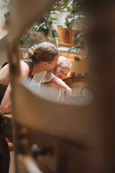 Orange County candid family photography of a mother kissing her smiling toddler while cooking together in a cozy, plant-filled kitchen, captured in warm natural light.
