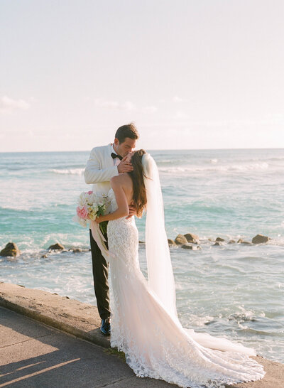 A bride in a lace wedding dress and groom in a white tux kiss  overlooking a Kauai beach.